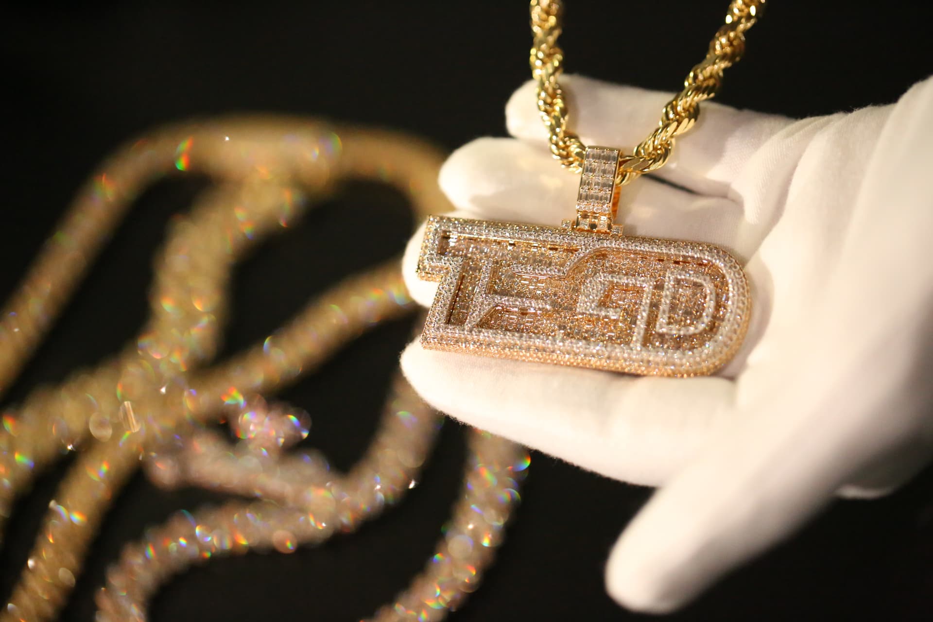 Diamond-encrusted letter pendant on a gold rope chain, held in a white-gloved hand